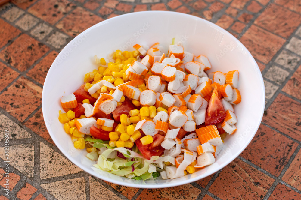 On a white plate, there's a multi-hued salad adorned with crab sticks, corn, and fresh green lettuce, enticing the appetite