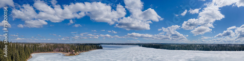 Panoramic view of a frozen lake that is surrounded by spruce trees. The sky is bright blue with puffy white spring clouds. A cabin can be seen on the shoreline.  © Craig Taylor Photo