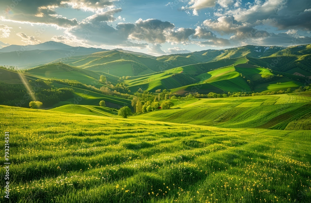   A lush green hillside, blanketed in grass, unfolds beneath a cloud-studded, blue-tinted sky Sunrays pierce the clouds