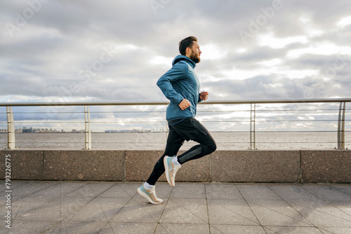 A determined athlete runs on a waterfront promenade, clouds above hinting at imminent weather. © muse studio