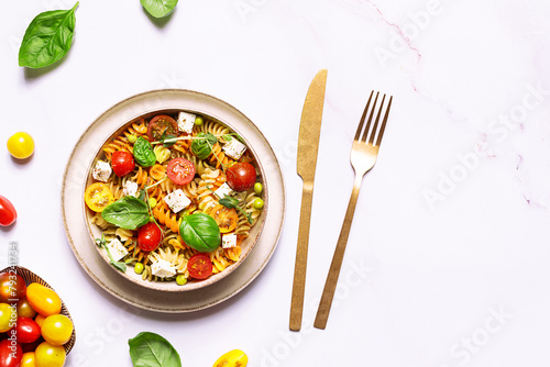 Colorful full grain fusilli pasta warm salad with feta cheese, cherry tomatoes, herbs, green pea and basil leaves on white stone background close up. Healthy diet food concept.