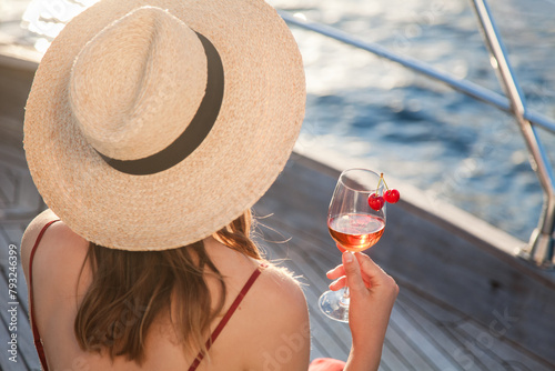 Woman drinking wine on yacht at sea. Summer vacation and travel at sunset. Elegant girl in straw hat relaxing, enjoying holidays and picnic with wineglass of alcohol-free beverage. Lifestyle moment
