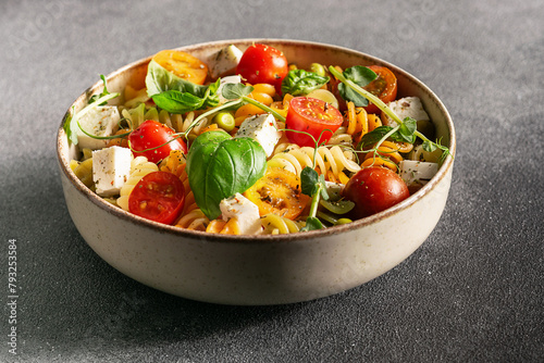 Colorful full grain fusilli pasta warm salad with feta cheese, cherry tomatoes, herbs, green pea and basil leaves on black stone background close up.