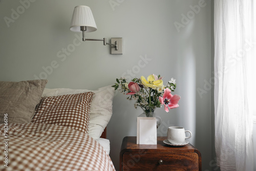 Spring interior still life. Blank vertical greeting card, invitation mockup. Elegant bedroom. Tulips, cherry tree blossoms bouquet in glass vase. Wooden night table. Cup of coffee. Checkered bedding.