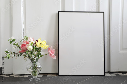 Spring still life scene. Tulips, cherry tree blossoms bouquet in glass vase. Blank black picture frame, poster mockup on grey tile floor. White wall background . Artisitic Easter decor, interior home. photo