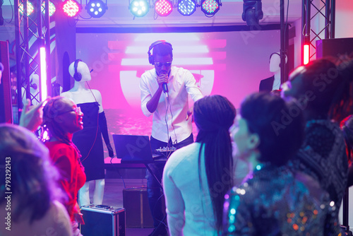 Quadro em tela Young man dj mixing music using controller and singing in microphone on club stage with spotlights