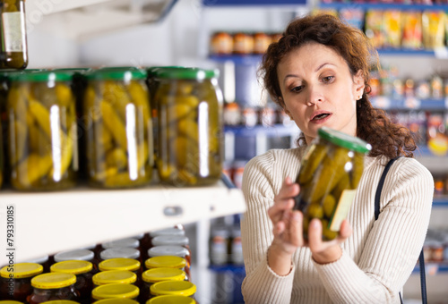 In Russian goods store  woman stand near showcase with canned vegetables  choose jar of pickled cucumbers. Buyer thinking about purchase  determine ratio of price and quality of products