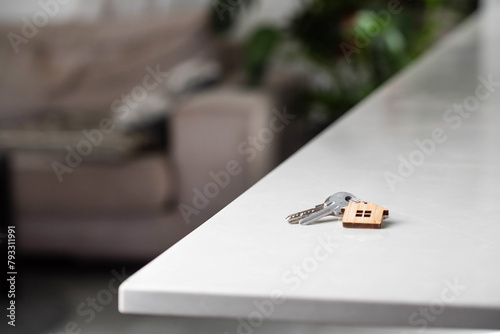 Keys with house shaped keychain on table on living room interior background. Concept of new homeownership, renting or leasing a property, and the process of purchasing a home, mortgage arrangements.