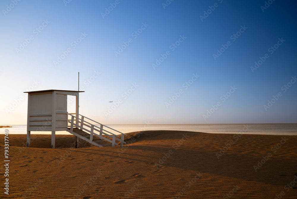 A small white building with a ladder leading up to it sits on a sandy beach. The sky is clear and blue, and there is a bird flying in the distance. The scene is peaceful and serene A small white building with a ladder leading up to it sits on a sandy beach. The sky is clear and blue, and there is a bird flying in the distance. The scene is peaceful and serene