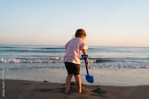 A young child gazes into the sea while holding a blue shovel on the beach, with the warm glow of sunset lighting the serene scene