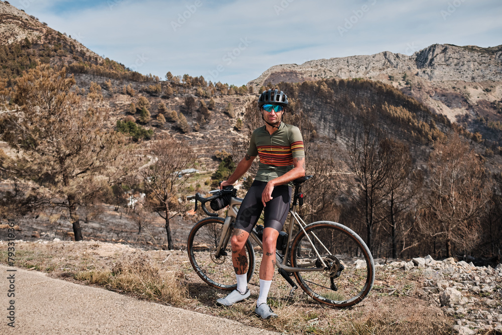 Portrait of a male cyclist in a helmet and cycling kit, standing near his bicycle on epic mountains background. Outdoor cycling. Healthy lifestyle concept. Coll de Rates pass in Spain