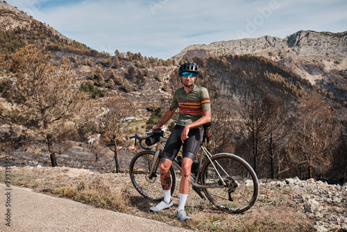 Portrait of a male cyclist in a helmet and cycling kit, standing near his bicycle on epic mountains background. Outdoor cycling. Healthy lifestyle concept. Coll de Rates pass in Spain