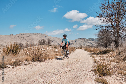 Cyclist riding gravel bike on gravel road in mountains with scenic view. Cyclist practicing on gravel road. Man cyclist wearing cycling kit and helmet. Alicante region in Spain.