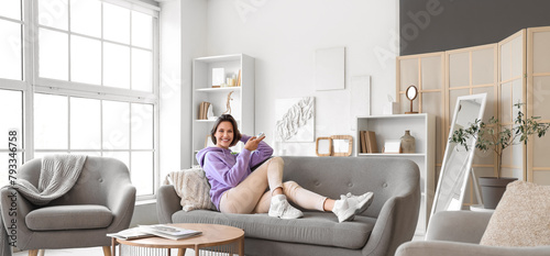 Фотография Young woman switching on air conditioner while sitting on sofa at home