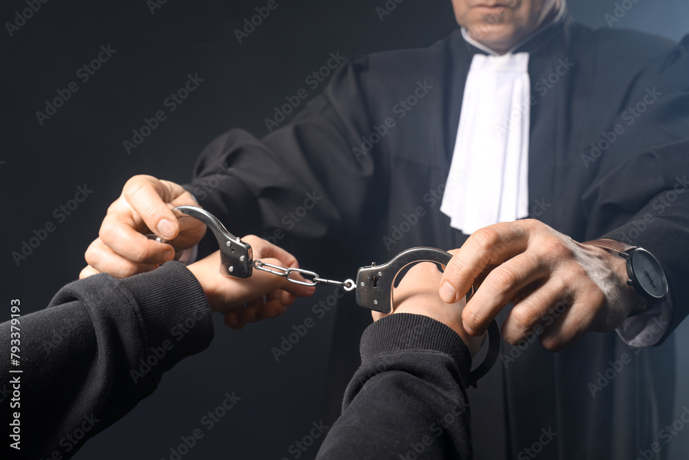Mature judge putting handcuffs on suspect against dark background, closeup