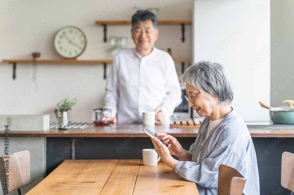 Obraz premium スマホを使う笑顔の高齢者女性とキッチンにいる高齢者男性 
