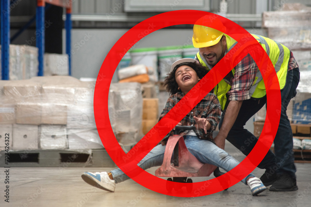 Dangerous, don't play in warehouses, prohibition sign superimposed  over the image of African child boy wearing a hard hat and reflective vest playing cart with male worker in warehouse
