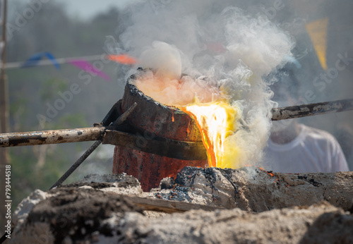 Obraz na plátně Blacksmith holding a hot crucible by the iron pliers from furnace and pouring the melting gold into the statue block
