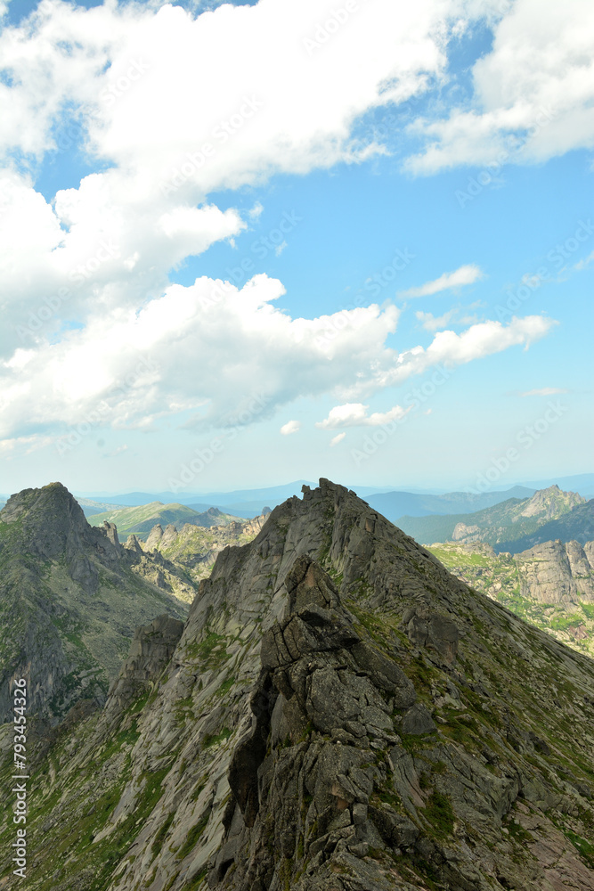 A narrow ridge of high rock with steep peaks in a chain of picturesque mountains under a cloudy sky on a clear summer day.