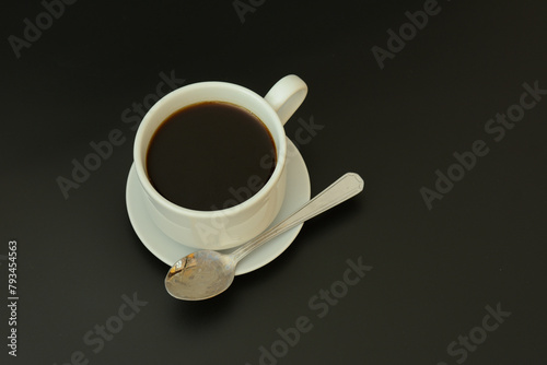 White ceramic cup of black coffee on a saucer with a small spoon on a black background.