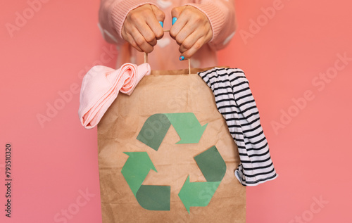 Paper bag with recycling sign with old clothes in hands against pink background.