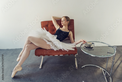 young ballerina in a tutu and pointe shoes is sitting on an armchair next to a table with a cup of coffee