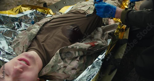 Confident male military man in a dark green uniform and with blue medical gloves cuts the T-shirt of a wounded soldier to stop bleeding during training and combat exercises providing medical care photo