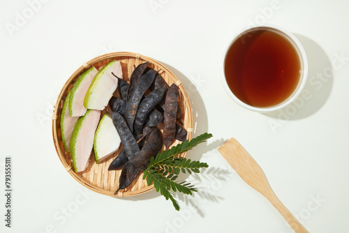 White background against a rattan tray with black locust seed pods and grapefruit peel on the left side and a bowl of brown liquid on the right. Copy space with view from above photo