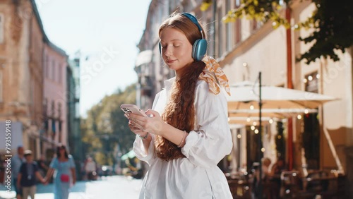 Happy relaxed overjoyed teenager girl in wireless headphones choosing, listening favorite energetic disco music in smartphone dancing outdoors. Child tourist walking passes by urban sunny city street photo