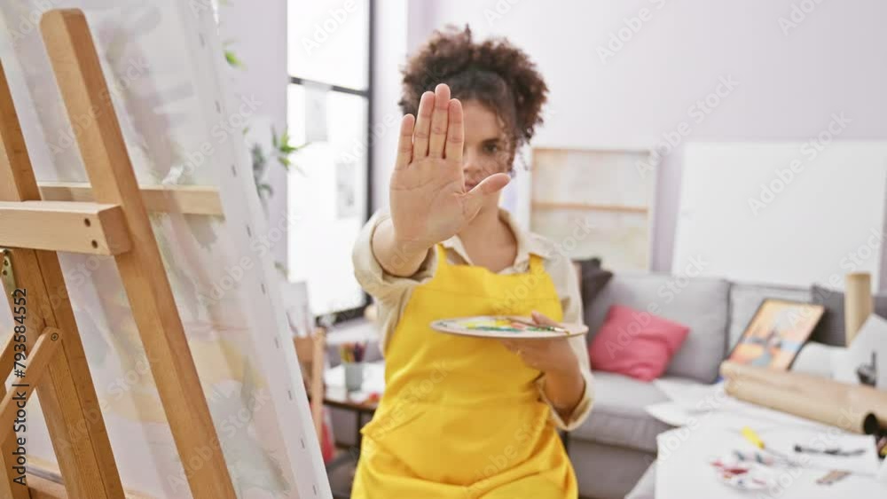 Hispanic curly hair woman artist sitting doing stop sing with palm of the hand. warning expression with negative and serious gesture on the face. at art studio Hispanic curly hair woman artist sitting doing stop sing with palm of the hand. warning expression with negative and serious gesture on the face. at art studio