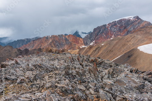 Awesome top view from rocky pass to sharp rocks on ridge of vivid colors against snowy peak in low clouds. Scenic landscape with colorful sheer crags and snow-capped mountain top under gray rainy sky.