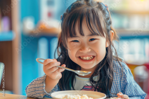 a happy young Asian girl eating at a table in a children's restaurant, holding a spoon and plate, with long hair bangs, focusing on her face, in natural light