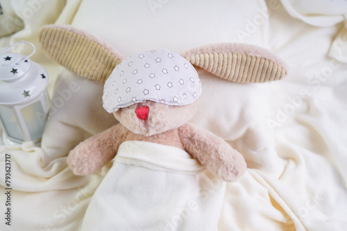 A stuffed rabbit sleeps with a mask over its eyes on a pillow under a white blanket