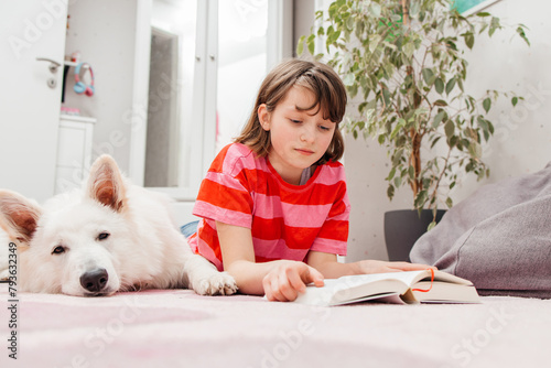 A teenage girl lies on the floor in her room and reads a book, next to her lies a dog, a white Swiss shepherd. Reading books as a hobby