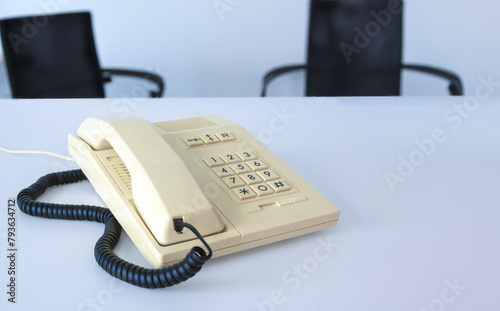 A white phone 80s - 90s with a cord hanging off it sits on a table in office