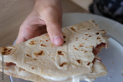 Hand Holding a Bitten Flat Bread Filled with Cheese and Ham on top of a Plate