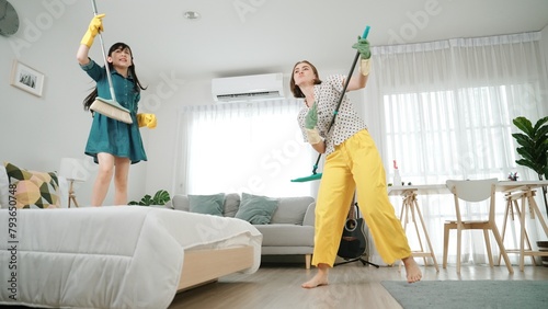Caucasian mom and asian child doing housework together and dancing to music. Attractive mother and daughter cleaning modern bedroom while sing along and jump to relaxing music. Lifestyle. Pedagogy.
