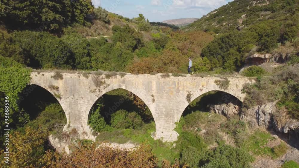 Adventurous Solo Hiker Walks Across an Ancient Aqueduct Bridge in Crete Greece. Stone Engineering Structure in Agricultural Farmland. Journey Down Beyond the Unknown. Adventurous Solo Hiker Walks Across an Ancient Aqueduct Bridge in Crete Greece. Stone Engineering Structure in Agricultural Farmland. Journey Down Beyond the Unknown.