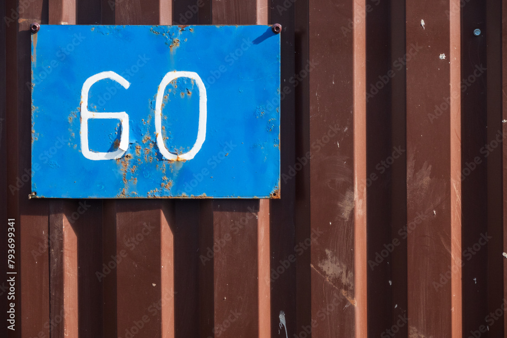 Fragment of a brown metal fence made of profiled iron. There is an old blue sign with the number 60. Background. Texture. Fragment of a brown metal fence made of profiled iron. There is an old blue sign with the number 60. Background. Texture.