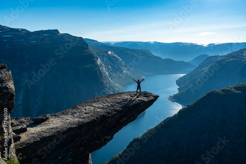 Trolltunga, Norway. Happy man tourist on the top of mountain's cliff edge named Trolltunga raising his hands up and watching Norwegian mountains. Adventure travel lifestyle emotional spectacular view.