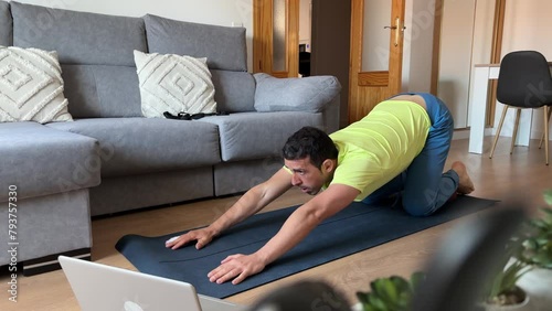 Man in quadruped position stretches arm and opposite leg with effort on yoga mat, accompanied by laptop and sleeping black cat in the couch. photo