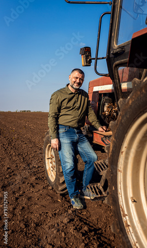 Portrait of satisfied farmer standing next to the tractor in the agricultural field.