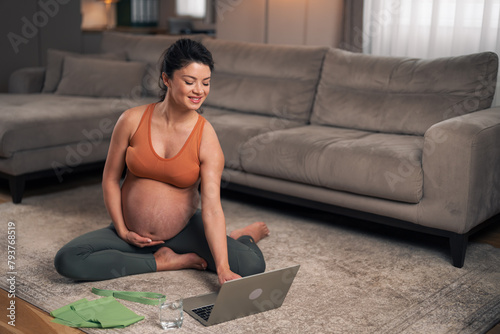 A motivated adult pregnant woman preparing to exercise at home by following a video tutorial on her laptop
