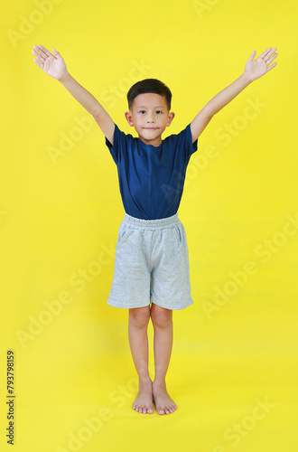 Asian boy child raised hands up. Kid standing and raise arms isolated on yellow background. Image full length.