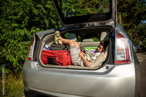 Cute little boy laying on the back of the bags and baggage in the car trunk ready to go on vacation with happy expression. Kid resting playing on smartphone