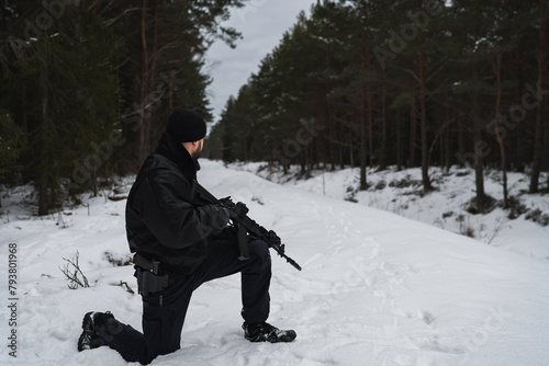 A military man in a black uniform with a rifle in his hands sits on his knee in the winter forest.