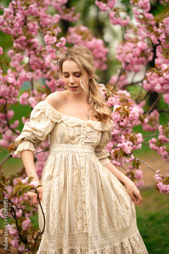 Pretty young blonde girl in vintage lace dress standing in spring park near pink blossom flowers. Tenderness romantic model posing 