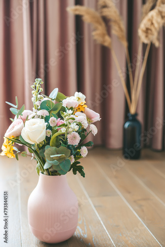 Assorted flowers in a textured pink vase stand out against elegant pink curtains and dried plants in a black vase