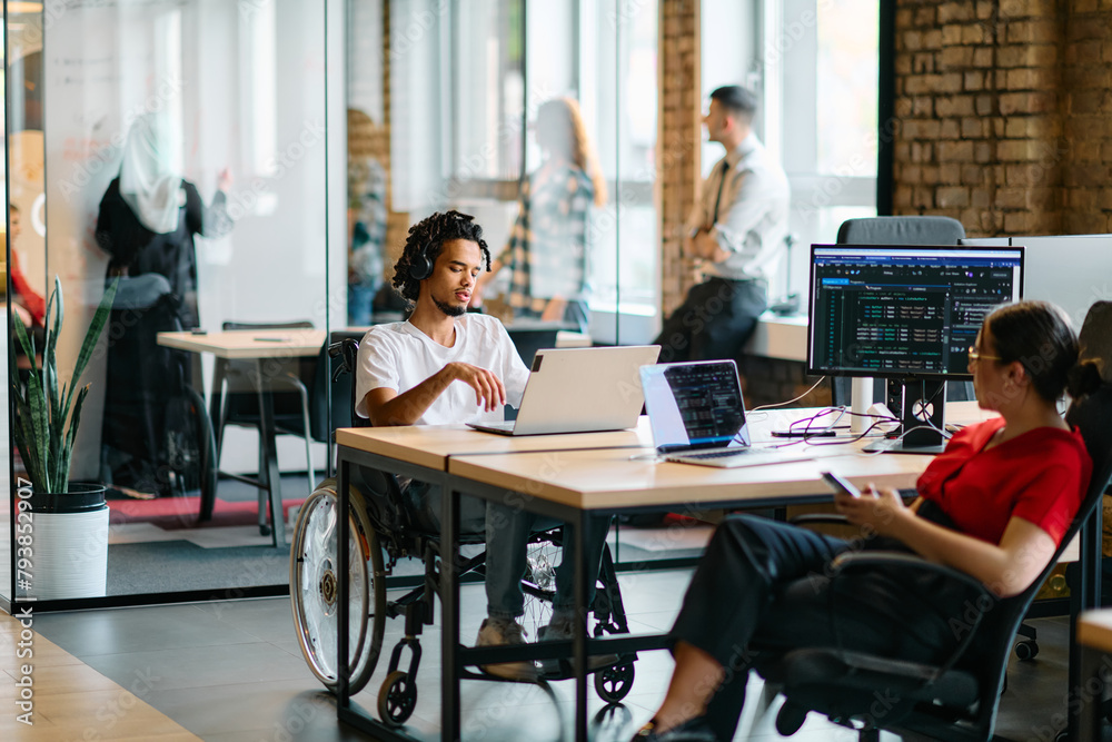 A young business group, including an African American businessman in a wheelchair, collaborates within a modern glass office, actively engaged around a computer and laptop, collectively solving