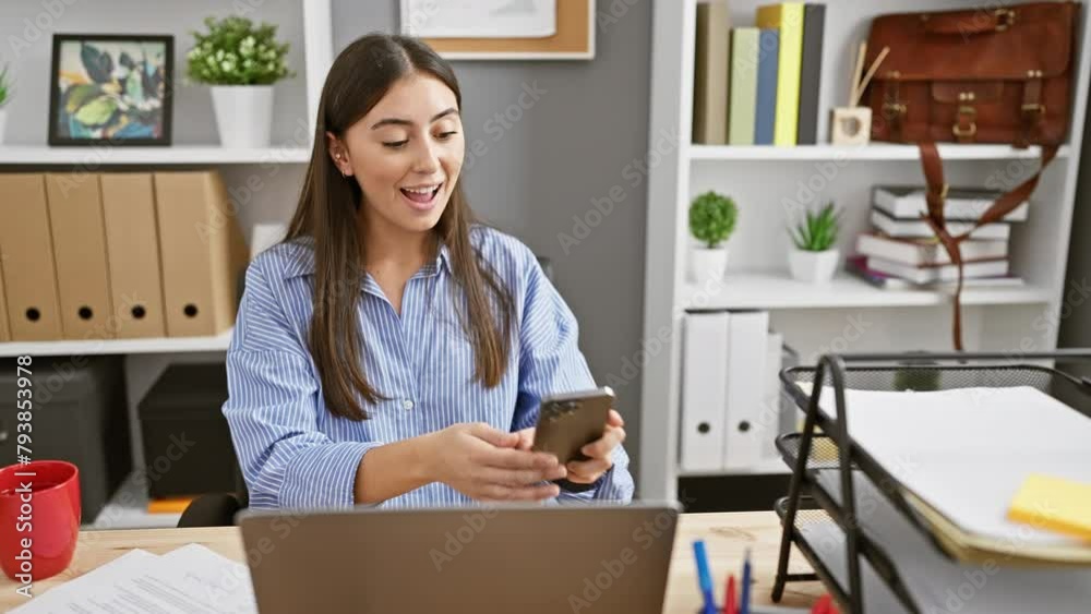 A young hispanic woman conducts a joyful video call from an organized indoor office, portraying an attractive, cheerful business environment.